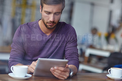 Buy stock photo Shot of a handsome young man using his tablet at a coffee shop