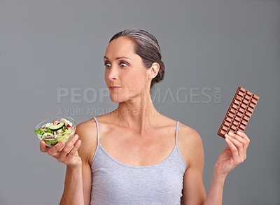 Buy stock photo Mature woman, salad bowl or chocolate with decision for choice, pick or selection in studio on a gray background. Female person, nutritionist or model with organic or mix vegetables and slab for diet