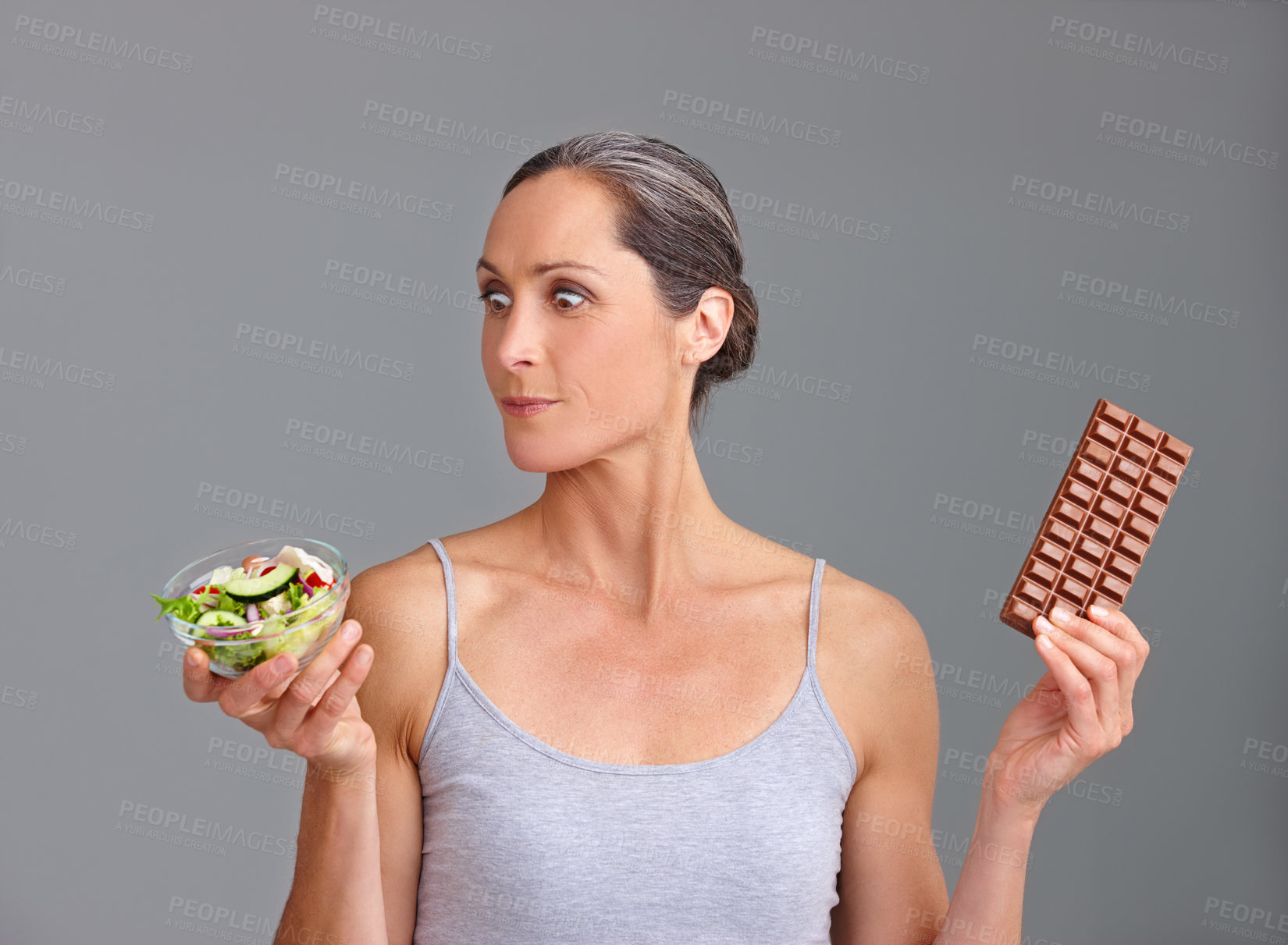 Buy stock photo Mature woman, salad bowl or chocolate with decision for choice, pick or selection in studio on a gray background. Female person, nutritionist or model with organic or mix vegetables and slab for diet