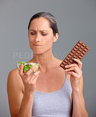 Buy stock photo Mature woman, salad bowl or chocolate with selection for diet, pick or choice in studio on a gray background. Female person, nutritionist or model with organic or mix vegetables and slab for decision