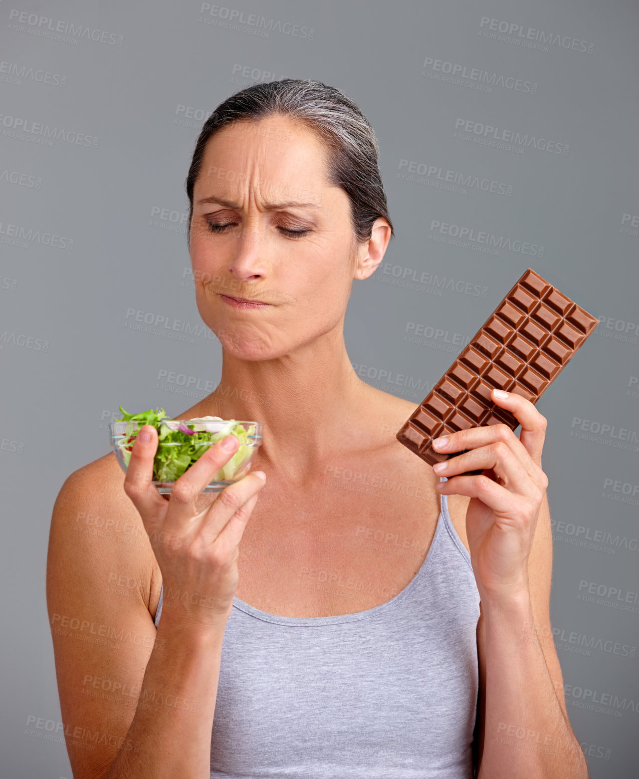 Buy stock photo Mature woman, salad bowl or chocolate with selection for diet, pick or choice in studio on a gray background. Female person, nutritionist or model with organic or mix vegetables and slab for decision