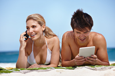 Buy stock photo Shot of a happy young couple using their tablet and cellphone at the beach