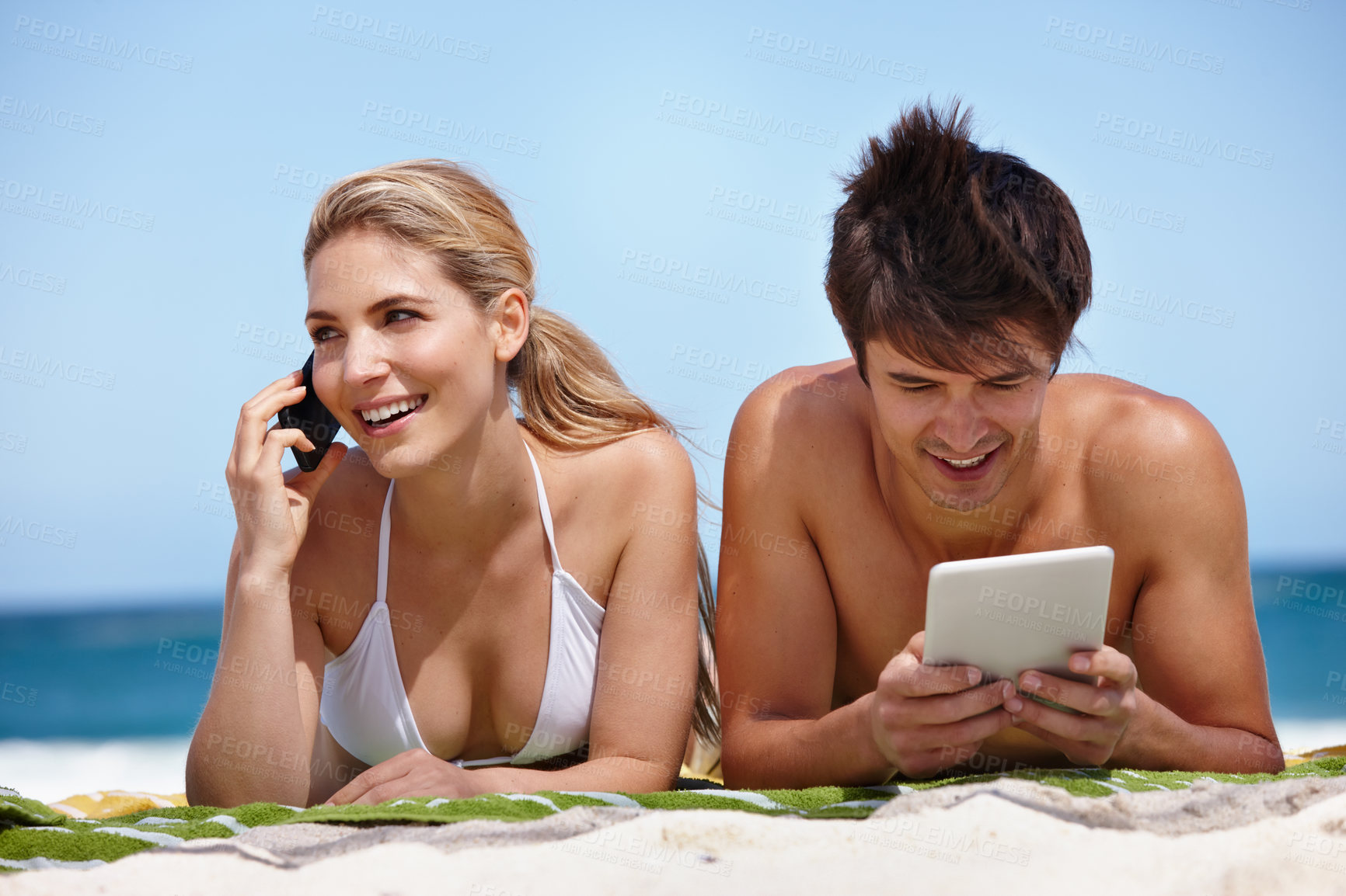 Buy stock photo Shot of a happy young couple using their tablet and cellphone at the beach