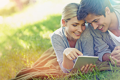 Buy stock photo Shot of a happy young couple lying on the grass and using a digital tablet together