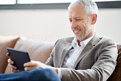 Buy stock photo Shot of a handsome mature man using his tablet while sitting on the sofa at home
