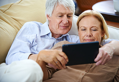 Buy stock photo Shot of a happy elderly couple watching something on a digital tablet while relaxing on their sofa