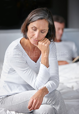 Buy stock photo Shot of a mature woman looking sad while sitting on her bed with her husband in the background