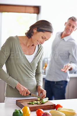Buy stock photo Shot of a mature woman chopping vegetables at a kitchen counter with her husband in the background
