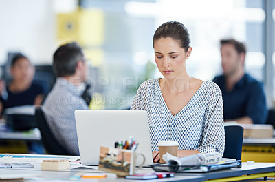 Buy stock photo Shot of a designer sitting at her desk working on a laptop with colleagues in the background
