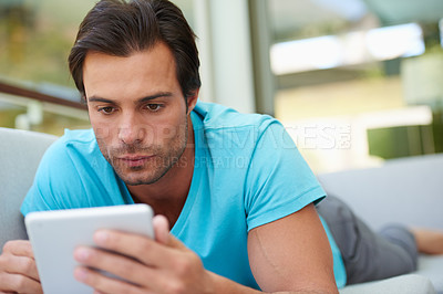 Buy stock photo Shot of a handsome man using his tablet while lying on the sofa at home