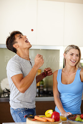 Buy stock photo Shot of a young man catching a grape in his mouth while his girlfriend prepares a meal