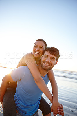 Buy stock photo Shot of a young man piggybacking his girlfriend on the beach at sunrise