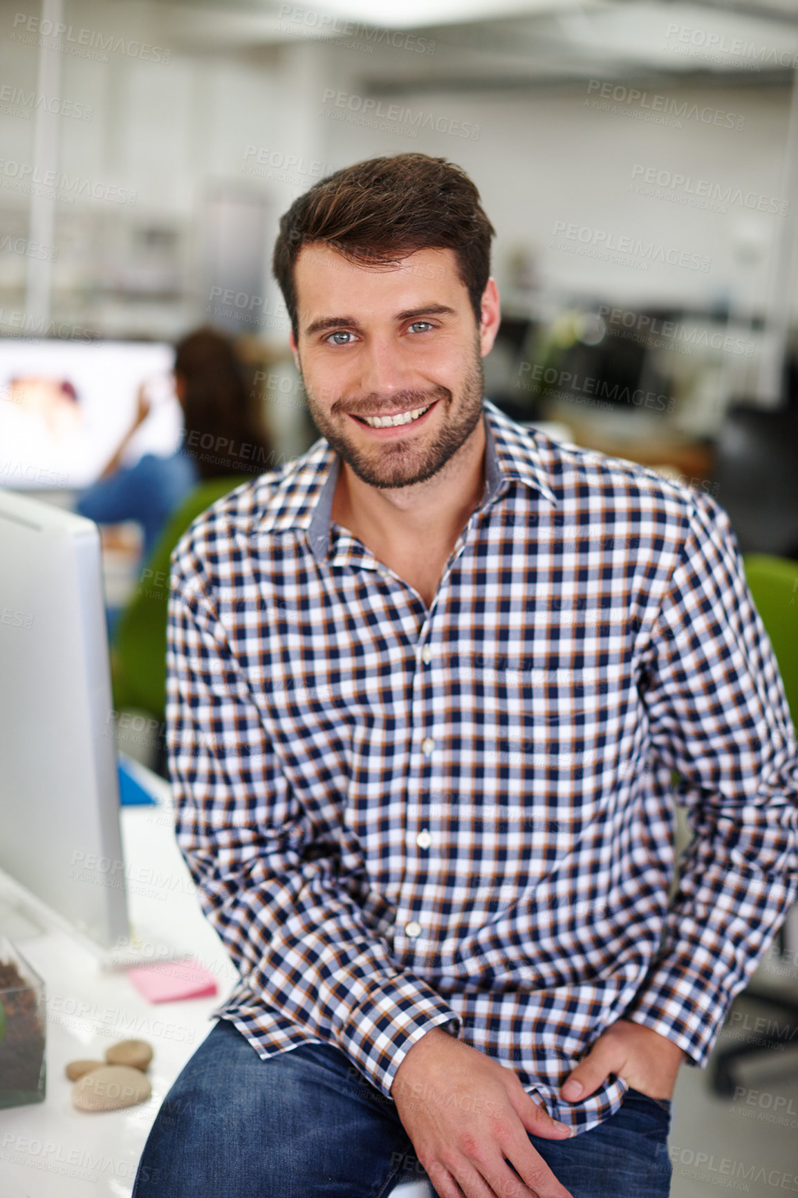 Buy stock photo Portrait of a handsome young designer sitting on his desk with a colleague in the background