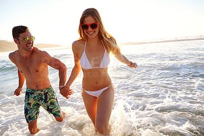 Buy stock photo Shot of a young couple in swimwear running through waves at the beach