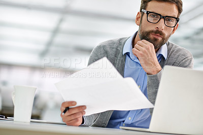 Buy stock photo A young businessman going over paperowrk while working on his laptop in the office
