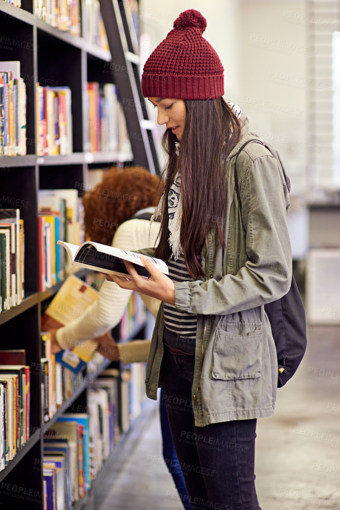 Buy stock photo Shot of a young woman studying from a book while standing by a library bookshelf