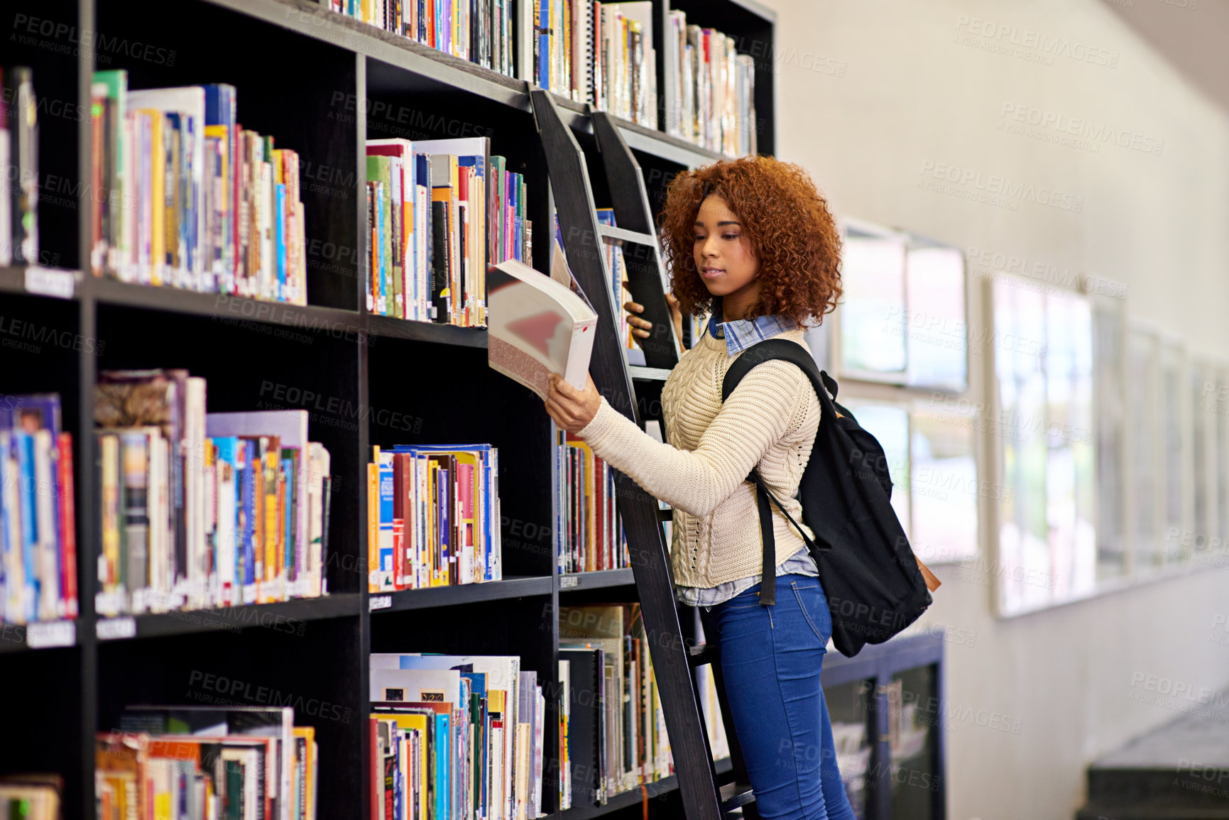 Buy stock photo Shot of a young woman studying from a book while standing by a library bookshelf