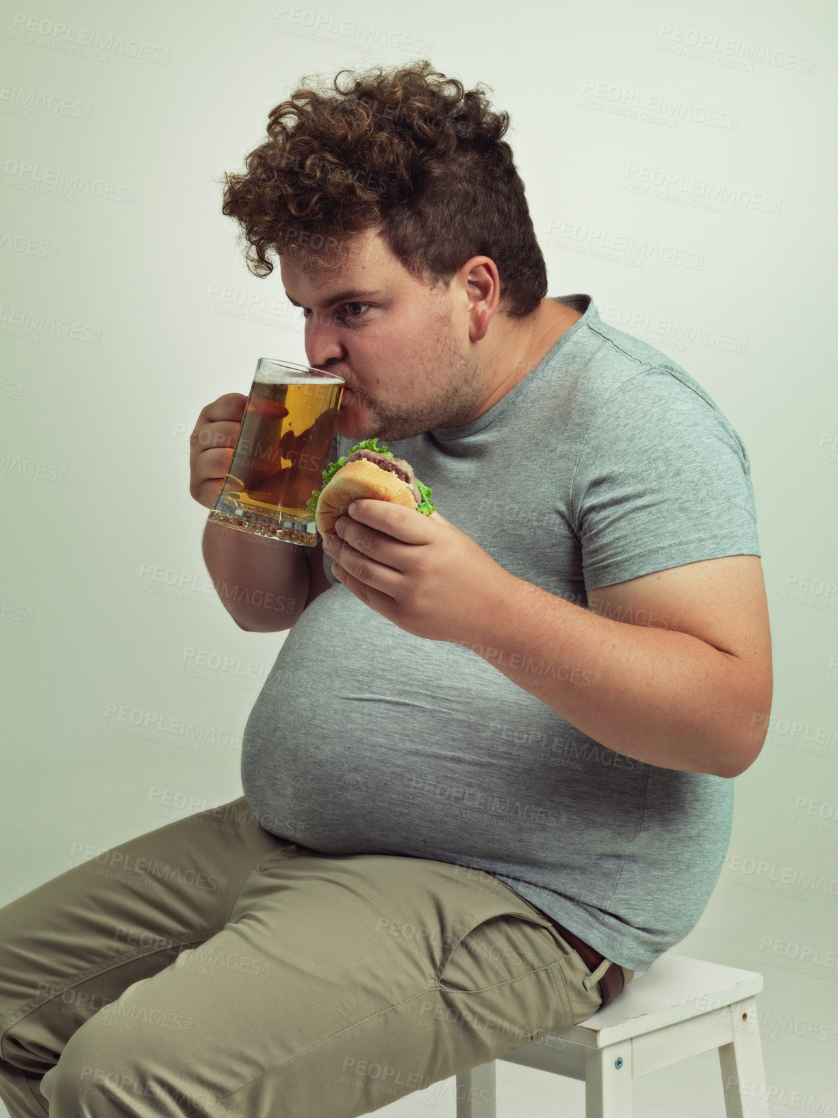 Buy stock photo Man, drink and beer with burger in studio for unhealthy diet, fast food and nutrition on chair. Plus size, hungry and male person with alcohol on stool for dinner, meal and beverage in glass