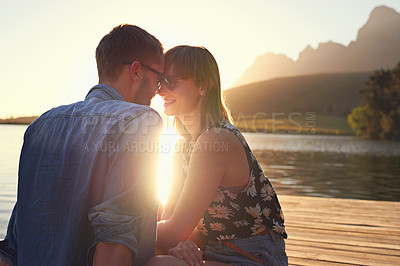 Buy stock photo Shot of an affectionate young couple sitting on a dock at sunset