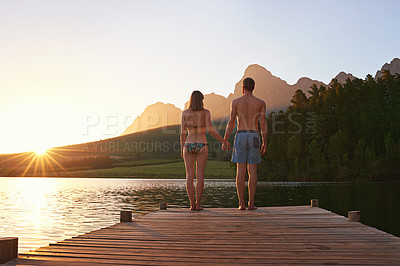 Buy stock photo Rearview shot of a young couple in swimsuits standing on a dock at sunset