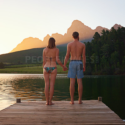 Buy stock photo Couple, holding hands and love with swimwear by lake with forest view, scenery and relax on summer adventure. Back, people and sunset on pier in countryside for romantic vacation and bonding together