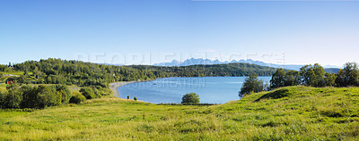 Buy stock photo Landscape of a lake with trees near a field. Green hills by the seaside with a blue sky in Norway. A calm lagoon near a vibrant wilderness against a bright cloudy horizon. Peaceful wild nature scene