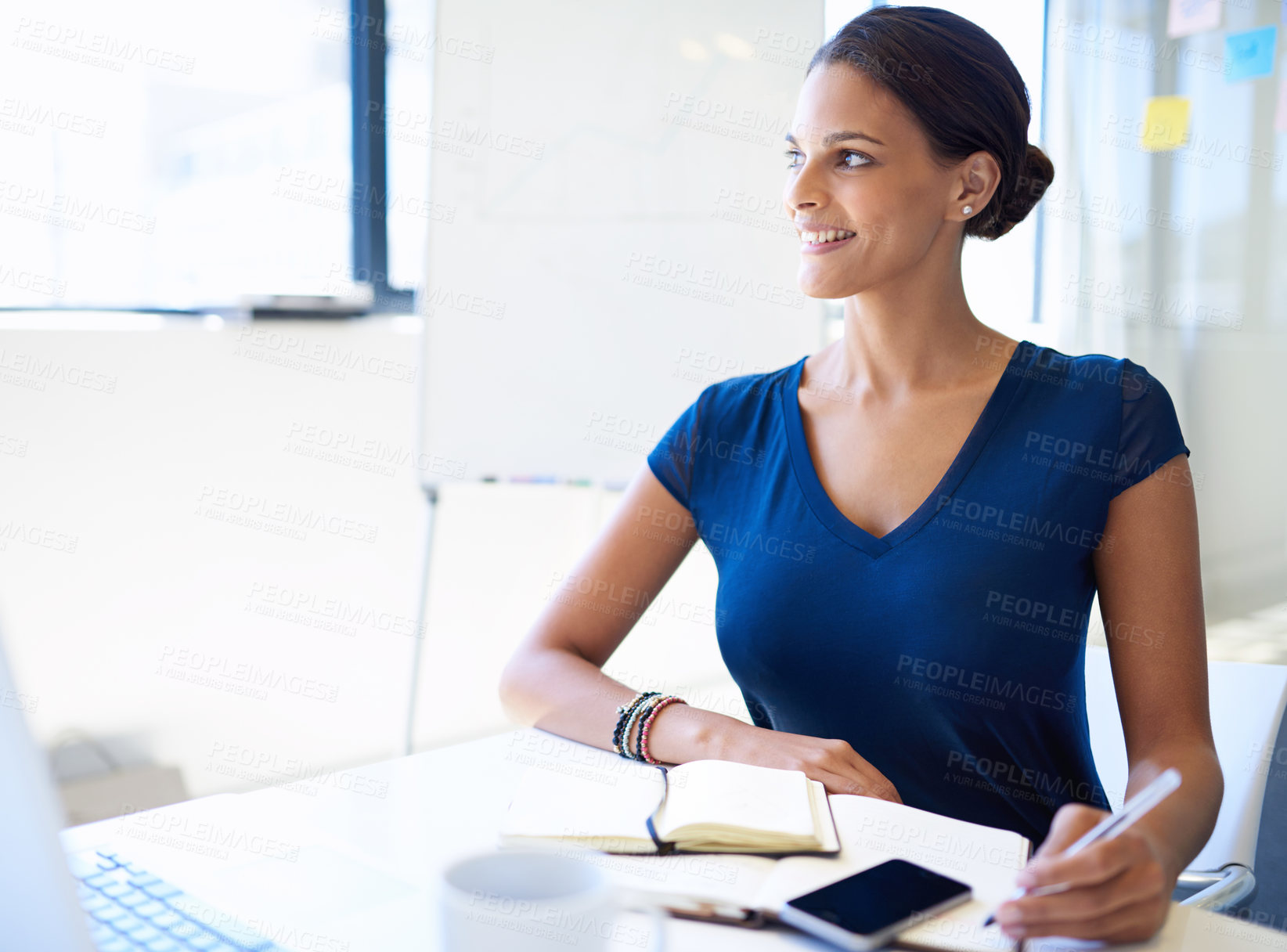 Buy stock photo A young businesswoman writing in a notebook while sitting with her laptop and cellphone