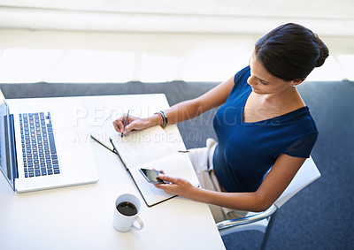 Buy stock photo A young businesswoman using her cellphone while sitting with her laptop and a notebook at her desk