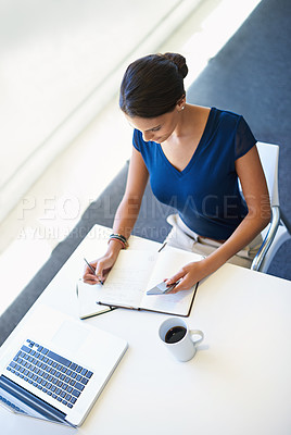 Buy stock photo A young businesswoman writing in a notebook while sitting with her laptop and cellphone