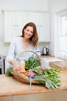Buy stock photo A beautiful woman stands in her kitchen looking at a basket full of fresh vegetables