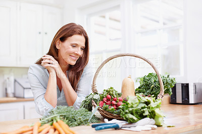 Buy stock photo A beautiful woman leans on her kitchen counter while looking at a basket of fresh vegetables