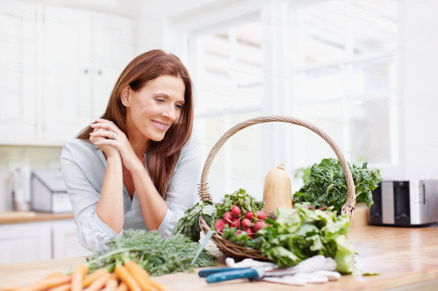 Buy stock photo A beautiful woman leans on her kitchen counter while looking at a basket of fresh vegetables