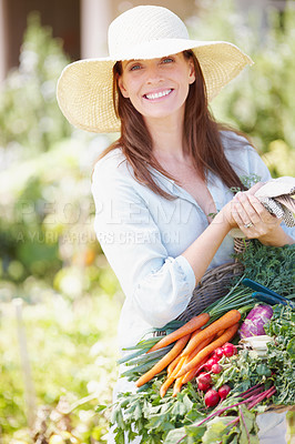 Buy stock photo A beautiful woman, wearing a straw hat, holds a basket of freshly picked vegetables while standing in her garden