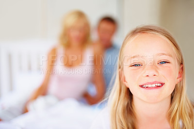 Buy stock photo Portrait, smile and a girl in the bedroom of a home with her parents in the background to relax on a weekend. Face, kids and a happy young child on a bed in an apartment to wake up in the morning