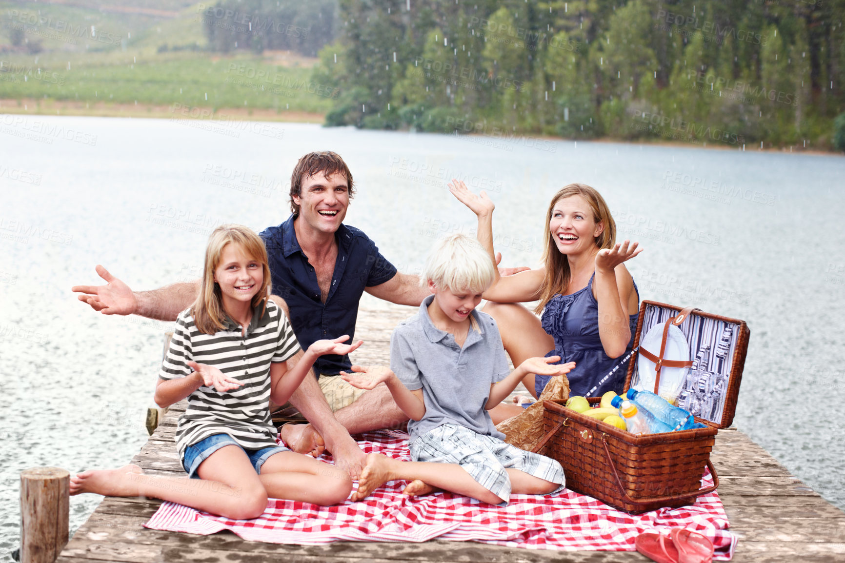 Buy stock photo Happy family, picnic and relax with rain outdoor at lake to celebrate, carefree or people laugh on water jetty. Mother, father or kids at river with food on vacation with hand gesture for wet weather