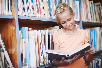 Buy stock photo Girl, library and smile for reading book at elementary school for literacy, knowledge and information. Student, kid and happy or satisfied with fairytale, novel and literature for brain development