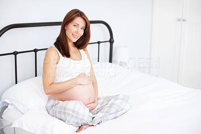 Buy stock photo Portrait of a beautiful pregnant woman holding her baby bump while sitting in bed