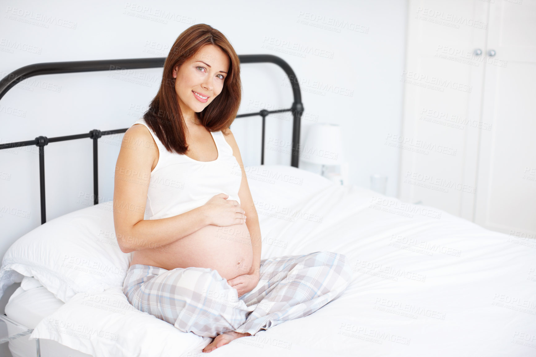 Buy stock photo Portrait of a beautiful pregnant woman holding her baby bump while sitting in bed