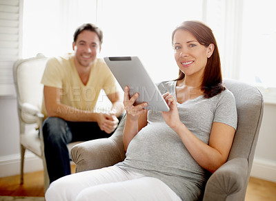 Buy stock photo A beautiful pregnant woman using a digital tablet while her husband sits in the background