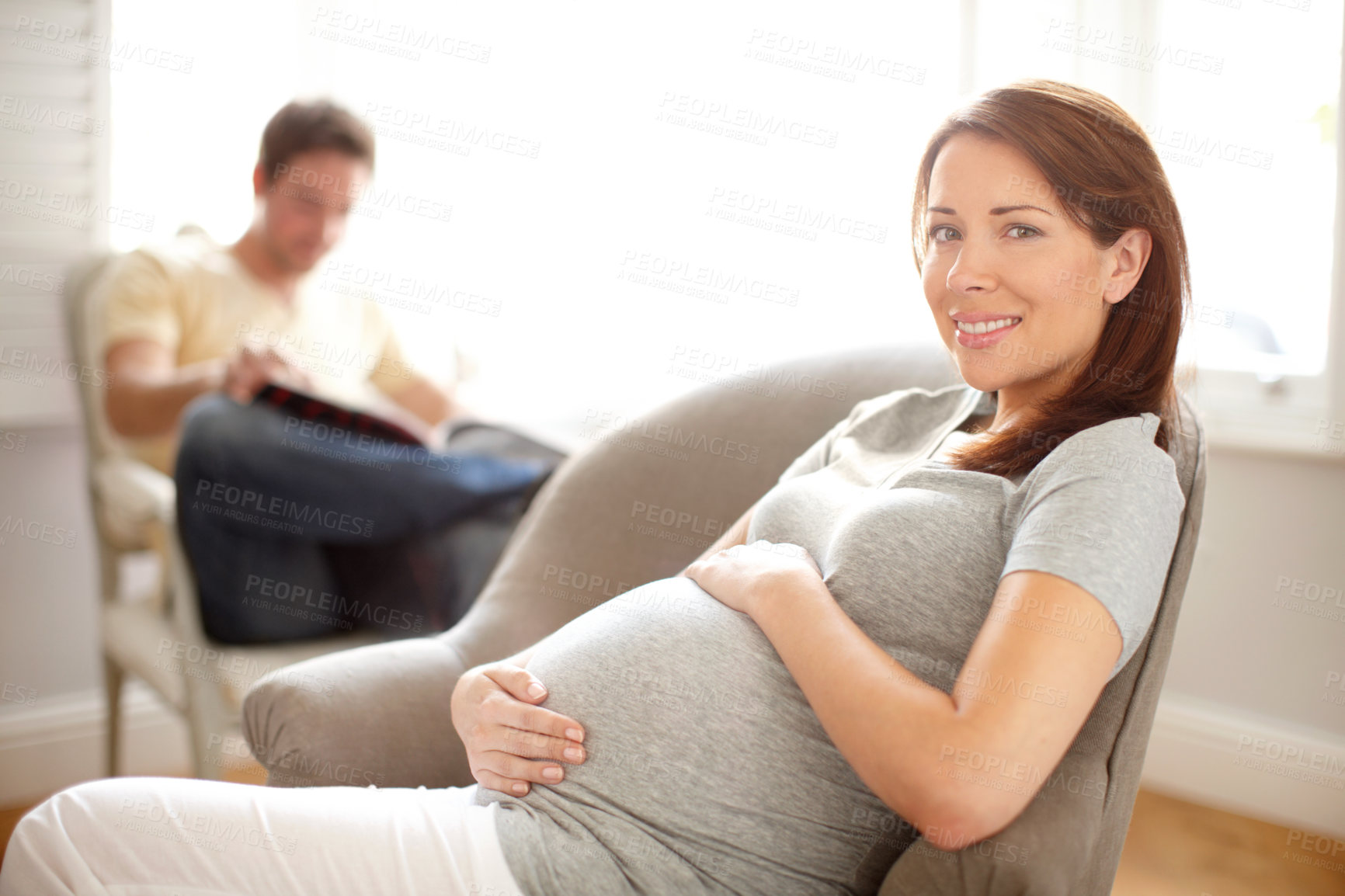 Buy stock photo Portrait of a beautiful pregnant woman holding her stomach while her husband sits in the background