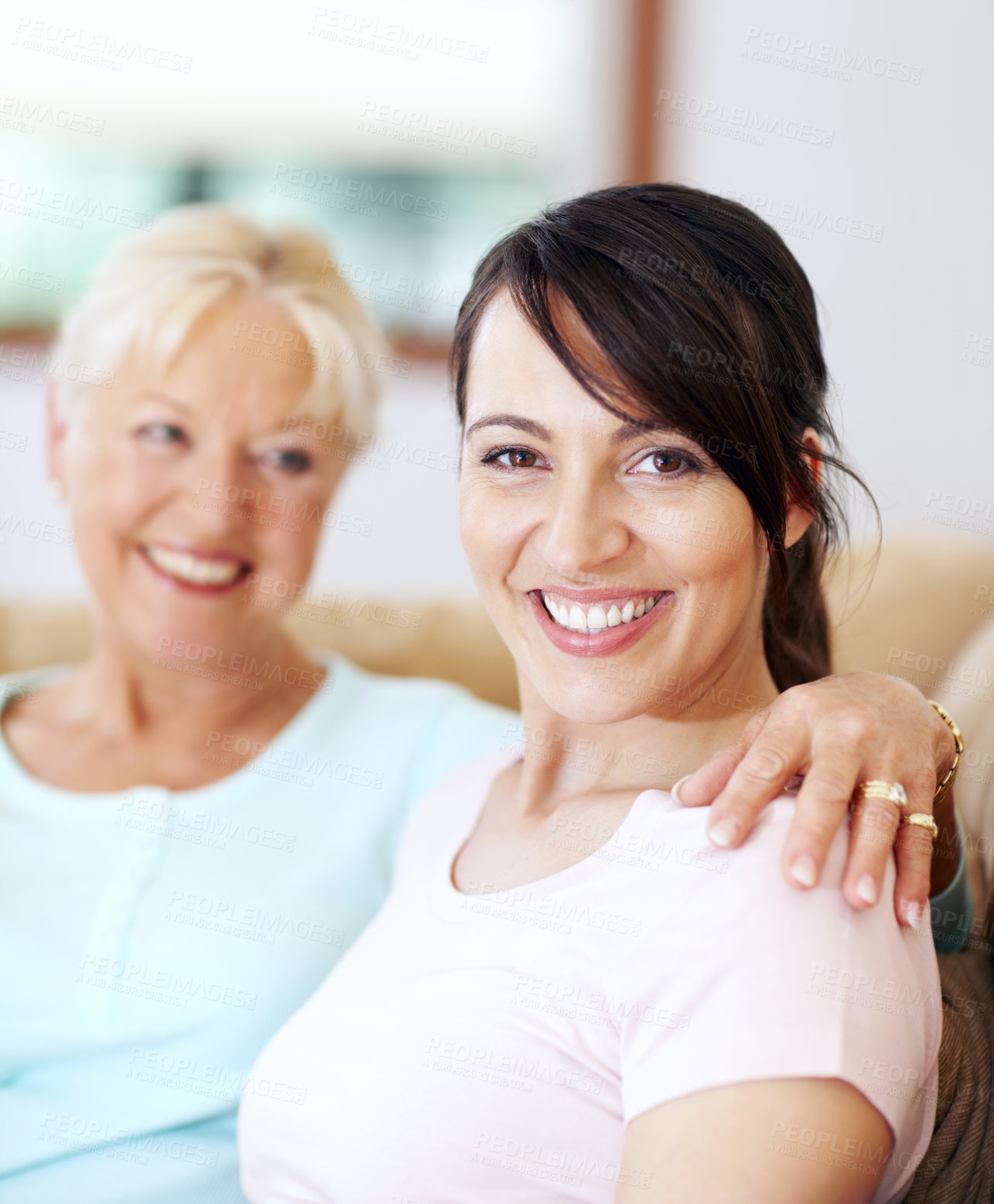 Buy stock photo Portrait of beautiful smiling woman sitting with her mother. Attractive brunette feeling happy on couch with her senior parent. Aged mother with her arm around her adult child spending time together 