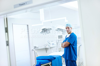 Buy stock photo A handsome male doctor standing next to an operating table with his arms crossed