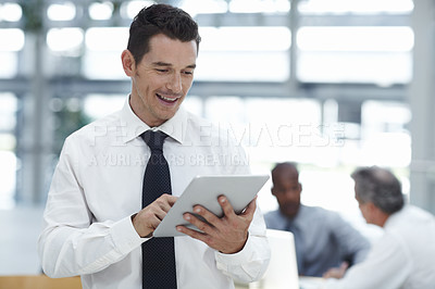 Buy stock photo A handsome businessman looking down at the touchpad in his hand and smiling with colleagues working in the background