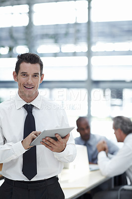 Buy stock photo A handsome businessman holding a touchpad smiling and looking at the camera with colleagues working in the background
