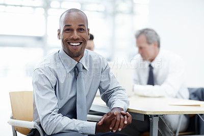 Buy stock photo A handsome African businessman sitting at a table and smiling with colleagues working in background