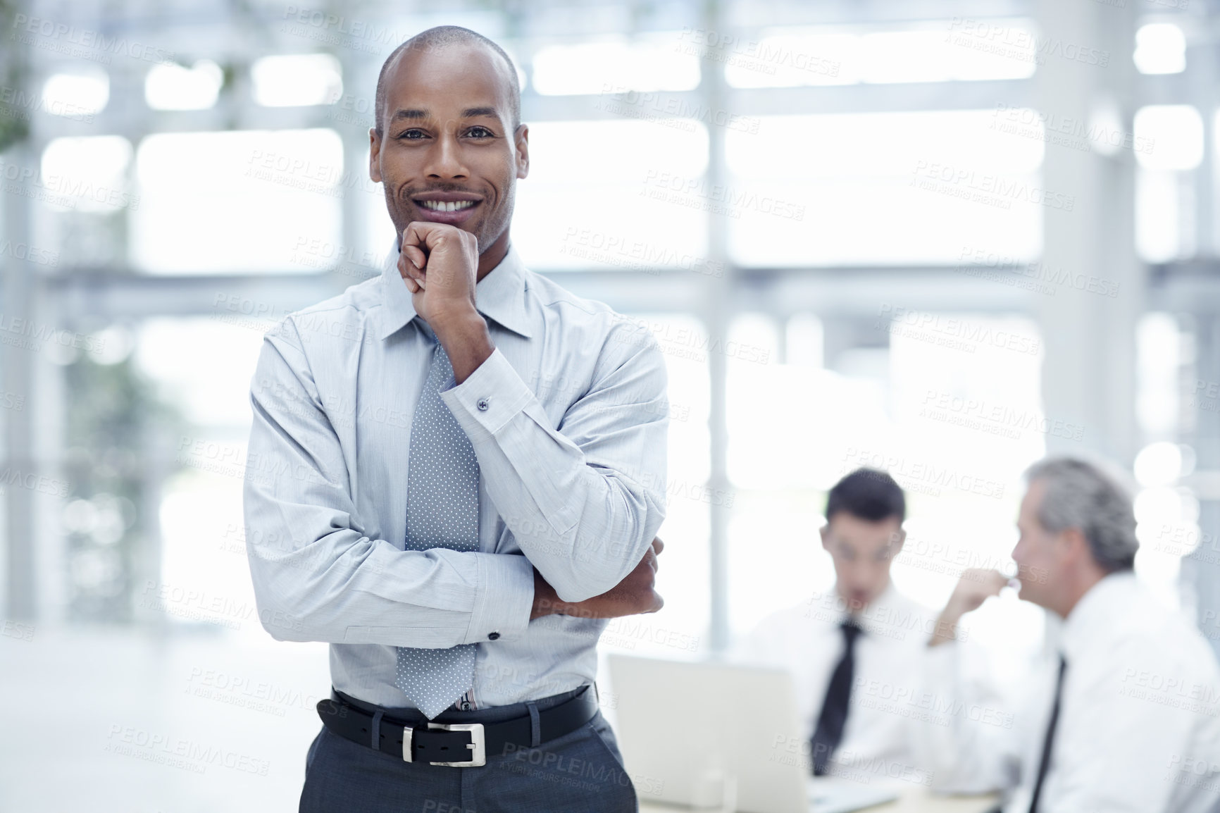 Buy stock photo A handsome African businessman stands with his chin rested on his hand, smiles and looks at the camera with colleagues in the background