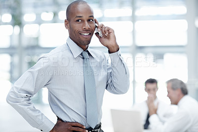 Buy stock photo A handsome African businessman, stands and talks on his cellphone with colleagues working in the background