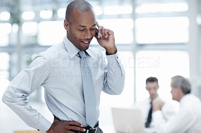 Buy stock photo A handsome African businessman talks on his cellphone and smiles, with colleagues working in the background