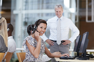 Buy stock photo A beautiful customer service representative sitting at her computer and talking on her headset with coworkers and manager in the background
