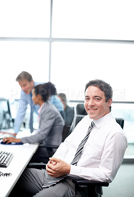 Buy stock photo Positive businessman sitting at his desk with his colleagues working in the background - portrait 